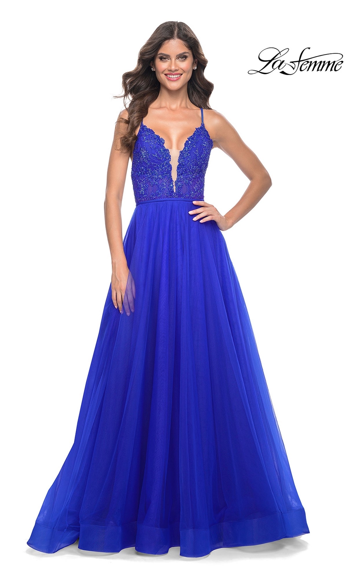 A woman stands smiling in the La Femme Deep V-Neck Long Tulle Prom Gown 32059, featuring a royal blue lace-bodice, thin straps, and a flowing A-line tulle skirt.